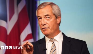 Reform UK leader Nigel Farage has grey hair parted to the side and is wearing a dark suit jacket, white shirt and striped tie