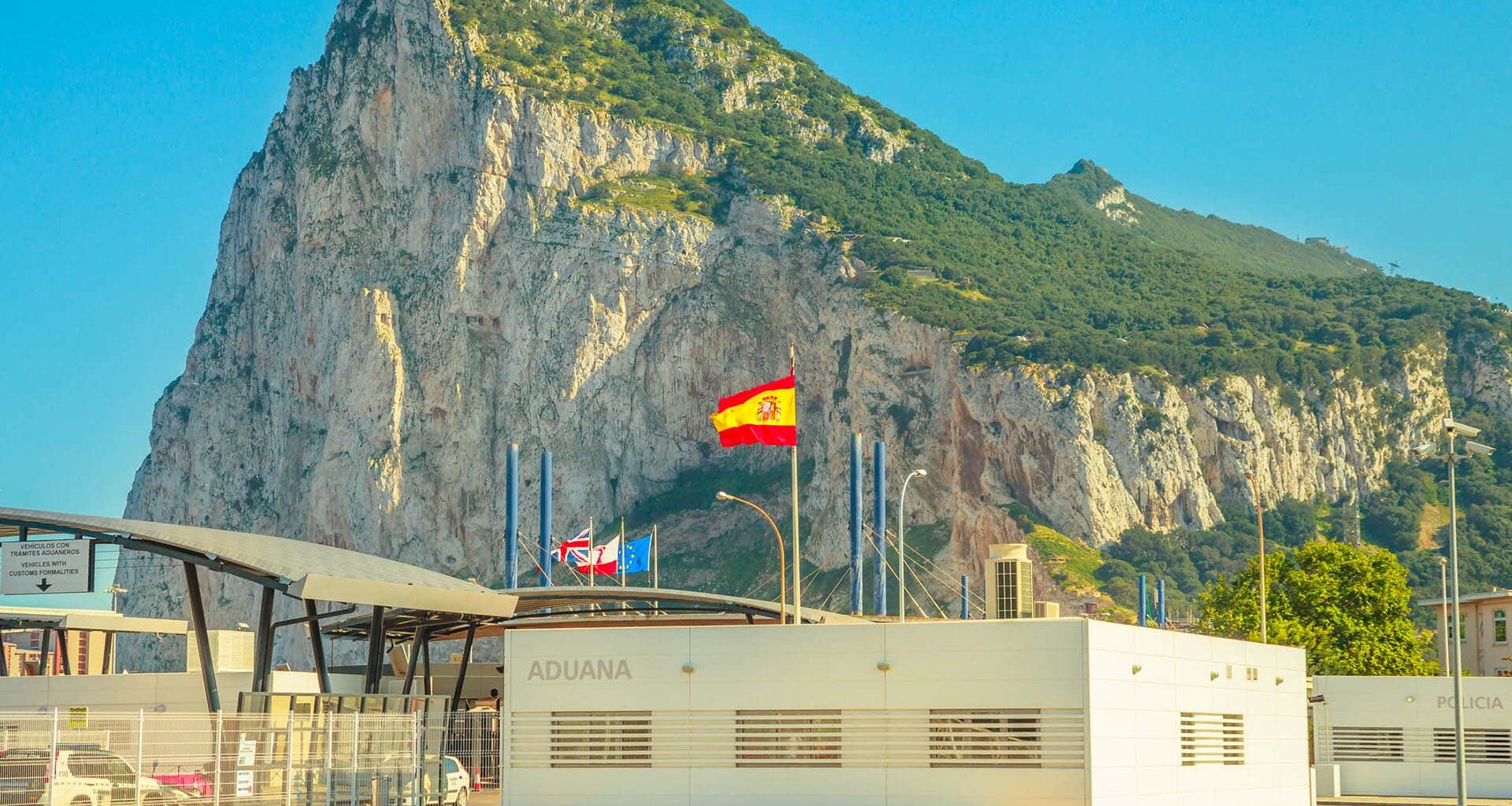 Border between Spain and Gibraltar with the Rock in the background, customs building in the foreground, and several flags flying on top (Spain, the UK, Gibraltar and the EU).