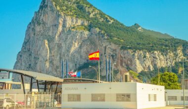 Border between Spain and Gibraltar with the Rock in the background, customs building in the foreground, and several flags flying on top (Spain, the UK, Gibraltar and the EU).