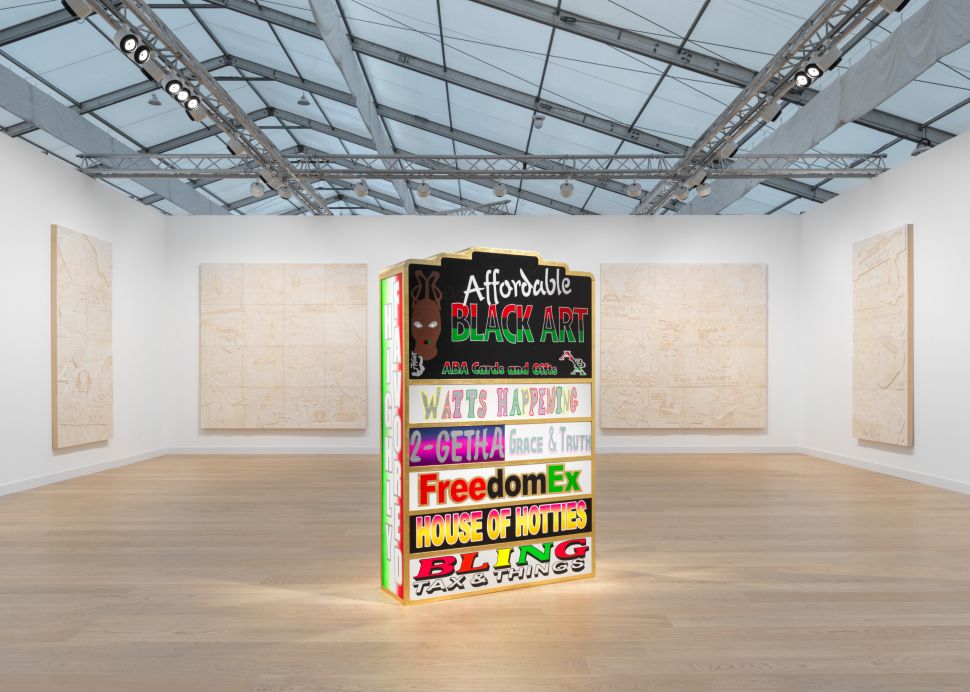 Colorful sculptural tower reading “Affordable Black Art” placed in the center of a booth surrounded by large monochrome carved panel works on every wall