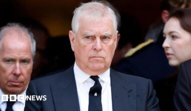 Prince Andrew, in dark blue double-breasted suit, white shirt and dark blue tie is standing in front of a man and a woman outside a church. The Prince has white hair and looks very serious.
