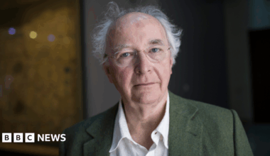 Sir Philip Pullman has wispy white hair and a bald crown. He is wearing a green blazer over a white shirt.