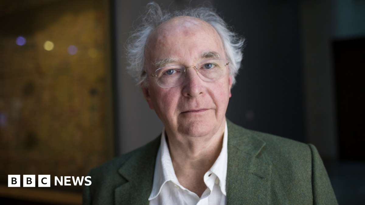 Sir Philip Pullman has wispy white hair and a bald crown. He is wearing a green blazer over a white shirt.