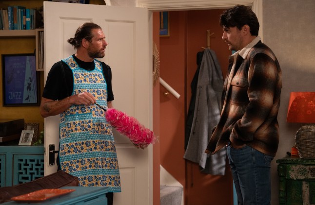 Kev in an apron holding a feather duster while talking to Mack in Emmerdale