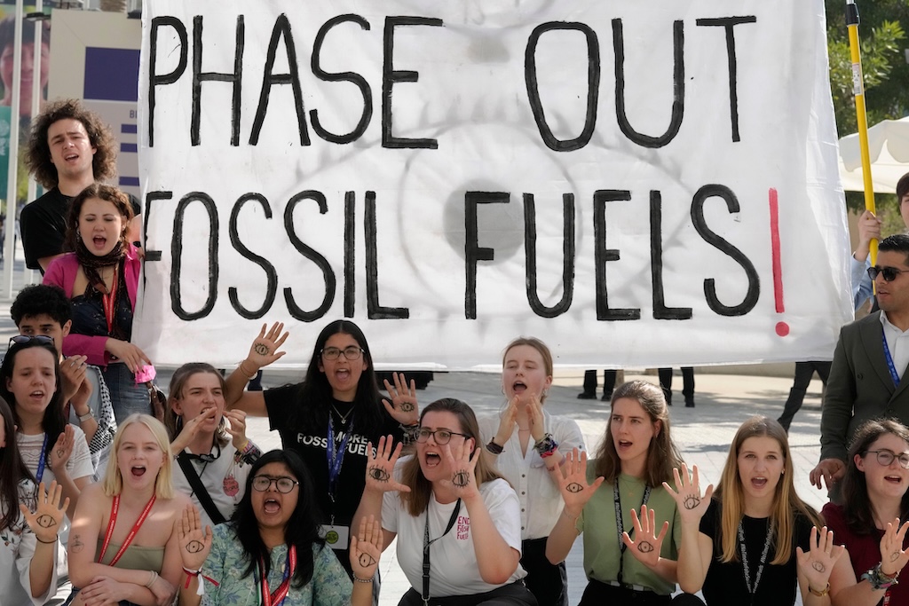 15 young people are pictured, some holding up their hands to show eyes drawn onto their palms. Some of them hold a large white banner with black writing that reads 'PHASE OUT FOSSIL FUELS!', the exclamation. point in red. They are all in the middle of saying or shouting something together.