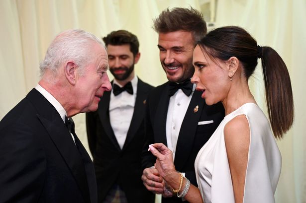 King Charles III, wearing a black suit and tie, talks to Victoria Beckham, wearing a white dress, while her husband David, wearing a black suit and tie, looks on