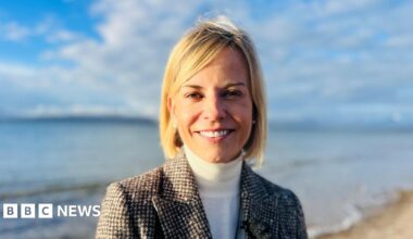 Susie Wolff, a woman with medium-length blonde hair, is standing on a beach somewhere rural. The sea and some distant land can be seen in the background. There is sand just behind her. She is wearing a black and white tweed checked jacket, white-polo-neck, and pearl ear-rings. She is smiling and it is sunny.