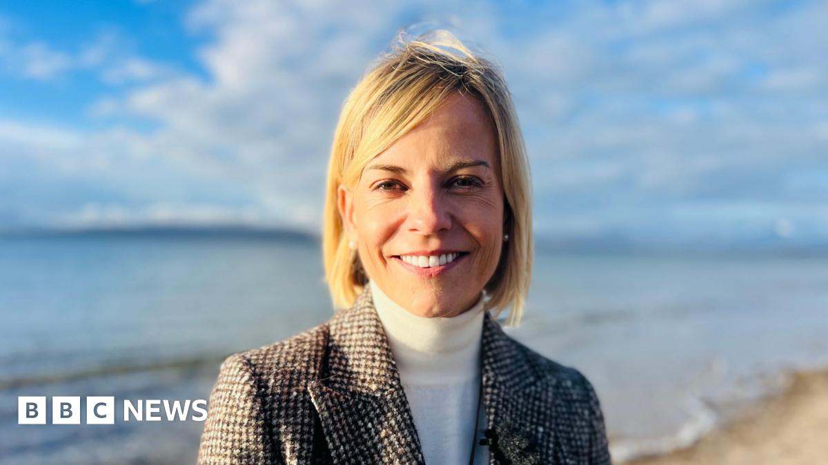 Susie Wolff, a woman with medium-length blonde hair, is standing on a beach somewhere rural. The sea and some distant land can be seen in the background. There is sand just behind her. She is wearing a black and white tweed checked jacket, white-polo-neck, and pearl ear-rings. She is smiling and it is sunny.