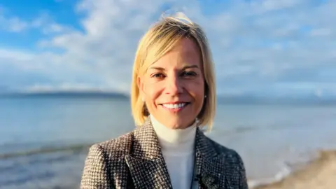 BBC Susie Wolff, a woman with medium-length blonde hair, is standing on a beach somewhere rural. The sea and some distant land can be seen in the background. There is sand just behind her. She is wearing a black and white tweed checked jacket, white-polo-neck, and pearl ear-rings. She is smiling and it is sunny.
