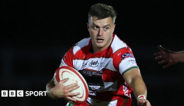 Ioan Hughes holds a rugby ball wearing a red and white hooped jersey