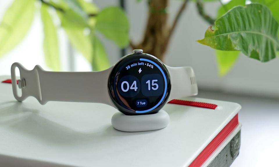 The Pixel Watch 4 laying on its charger, showing the time is 4:15PM and it's 54 percent charged. The screen also says there is 30 minutes left to fully charge the watch and a ring around its display shows charging progress.