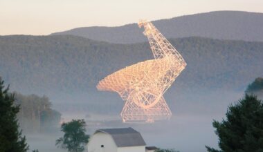 A large radiotelescope dish, white but lit by golden hour light, against a background of misty forested mountains.