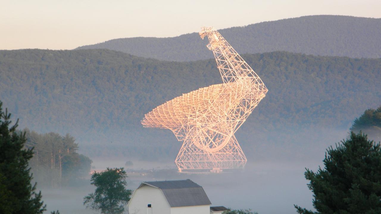 A large radiotelescope dish, white but lit by golden hour light, against a background of misty forested mountains.