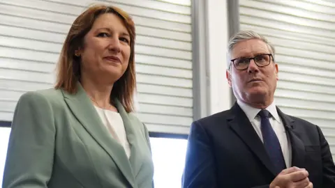 PA Media An image of Rachel Reeves on the left in a grey suit, and Keir Starmer on the right in a black suit, stood in front of windows with closed blinds, during a visit to Horiba Mira in Nuneaton in June 2025.