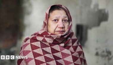 Nicola Wheatley is sitting on a  rock in a green space with her children on her lap. She and Oscar are smiling at the camera and baby Ffion is wearing a yellow summer hat and dungarees.