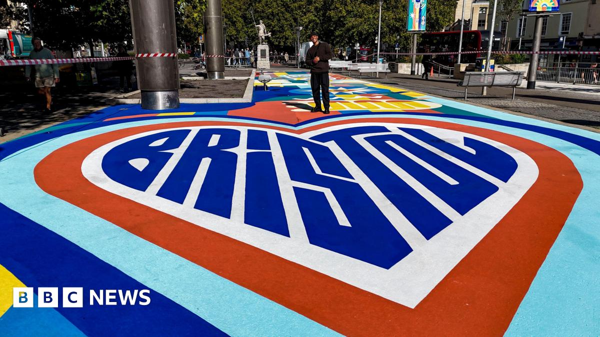 A close-in view of part of a new artwork in Bristol city centre. The art contains a giant heart shape in different colours, those being light blue, dark blue and red. Inside the heart is the word "Bristol" in large blue letters which are stylised to also be in the shape of a heart. At the top of the heart stands the artist who made it. He is wearing an all-black outfit