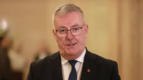 PA Media Mike Nesbitt wears a black suit and a white shirt with a navy tie. He has short white hair and wears glasses. 