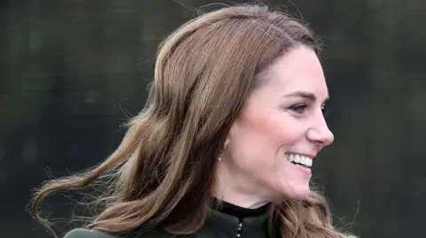 Chris Jackson/PA Wire A close-up of the Princess of Wales smiling.  She has long, wavy brown hair and is wearing a dark olive green coat fastened to the neck over a black top.