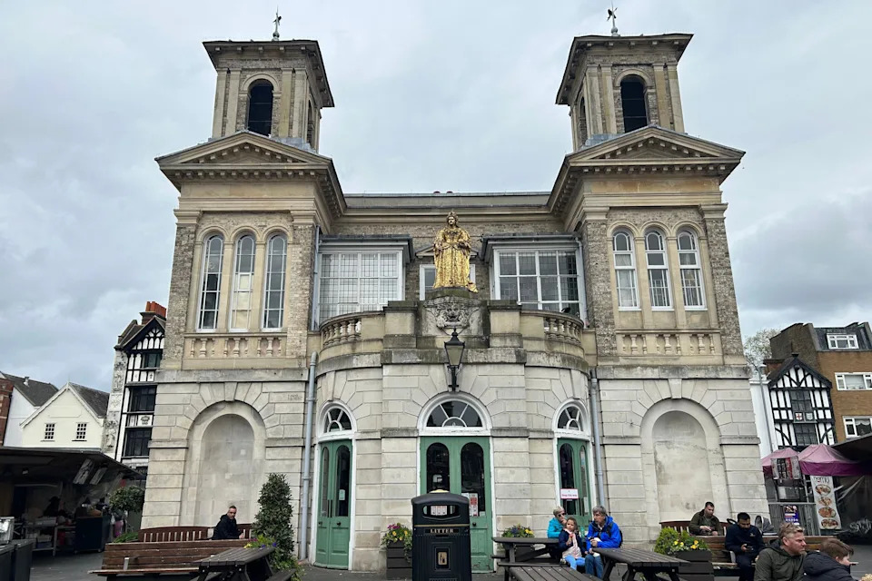An old, white brick building with three light green doors at the front. There is a golden statue above the doors, and a bin and benches in front of the building. This building is currently unoccupied.