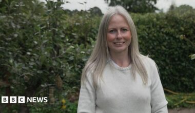 Emily, wearing a cream jumper, smiles as she stands in front of hedges and a tree with a bird feeder hanging in its branches
