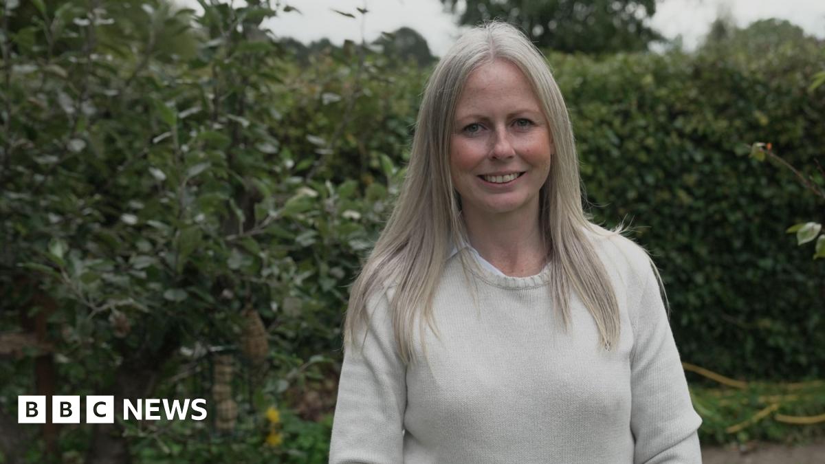 Emily, wearing a cream jumper, smiles as she stands in front of hedges and a tree with a bird feeder hanging in its branches