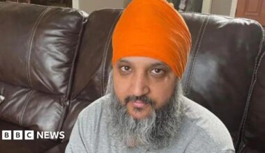 Paramjit Singh wearing an orange turban and grey shirt is sitting on a brown sofa.