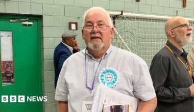 Andy Osborn stands in a counting hall. He is looking directly at the camera and is wearing a blue and white striped shirt with a blue and white Reform UK rosette pinned onto it. He has a purple lanyard around his neck and is carrying a set of papers. Behind him is a green door and a football goal is hanging up on the wall behind him.