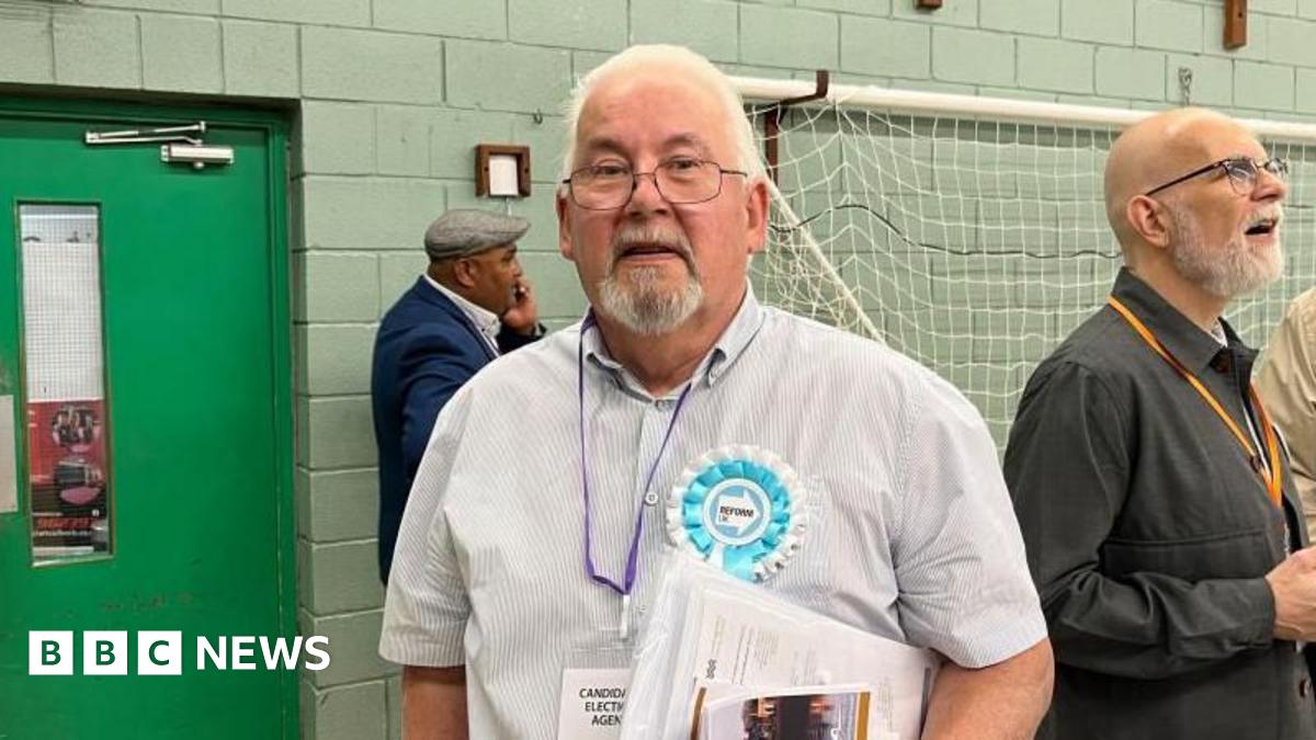 Andy Osborn stands in a counting hall. He is looking directly at the camera and is wearing a blue and white striped shirt with a blue and white Reform UK rosette pinned onto it. He has a purple lanyard around his neck and is carrying a set of papers. Behind him is a green door and a football goal is hanging up on the wall behind him.