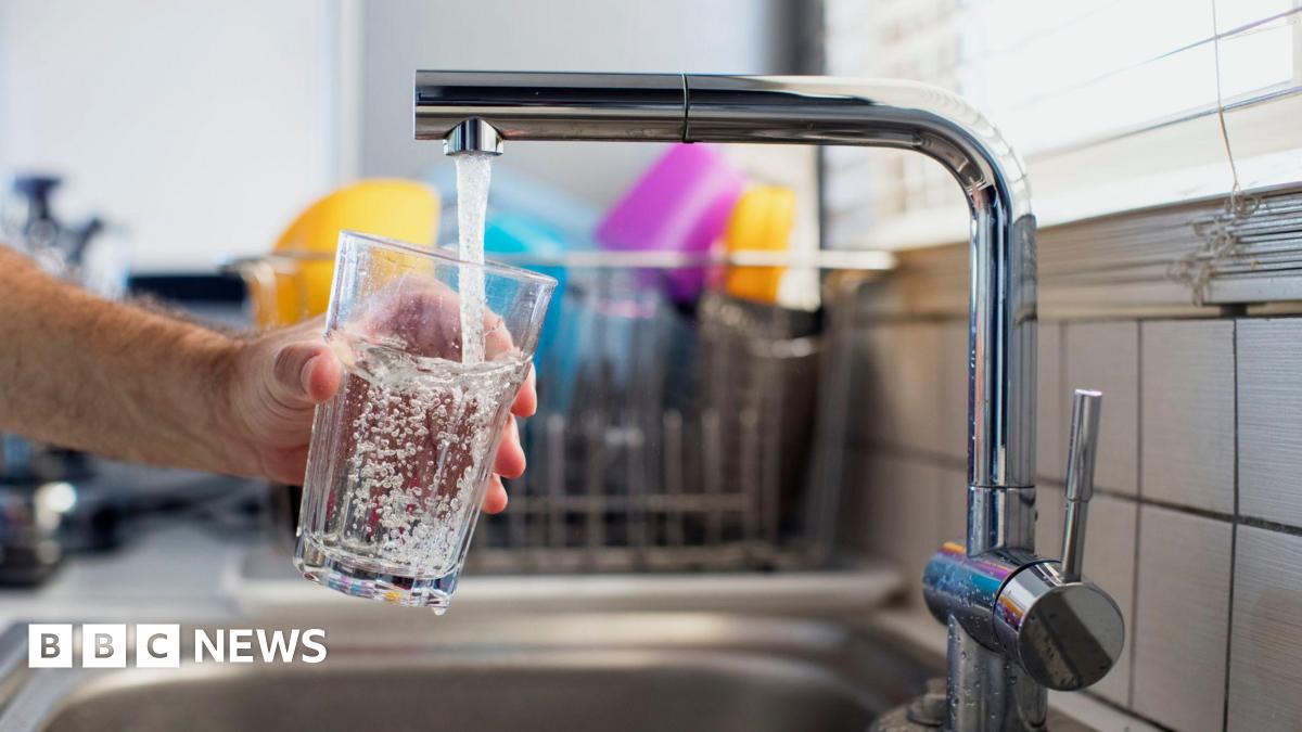 A person holding a clear glass under a running stainless steel tap, filling it with water. Behind the sink, a dish rack sits near a window with horizontal blinds.