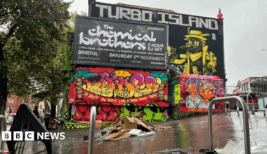 Photo of Turbo Island area. A building with graffiti and a black and white billboard. Park cars surrounding the area with bike racks. In the middle of the small area there is burnt debris and cardboard in a pile.