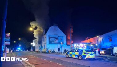 A police car is seen on a road in front of a building with smoke coming from the house. It is night time and the street lights are on.