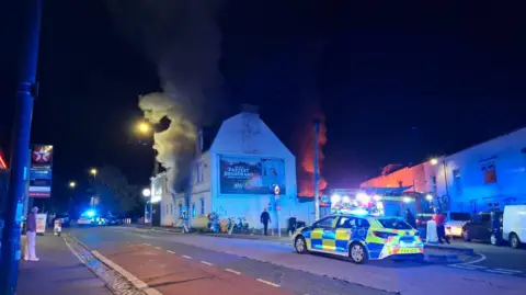 BBC A police car is seen on a road in front of a building with smoke coming from the house. It is night time and the street lights are on.