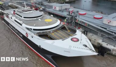 The fastcraft Manannan Steam Packet Company vessel in white and black, is docked alongside a long building with the Manx triskelion displayed on the left, right and centre of the building. People in high vis jackets can been seen on an elevated walkway connecting the ship to the land.