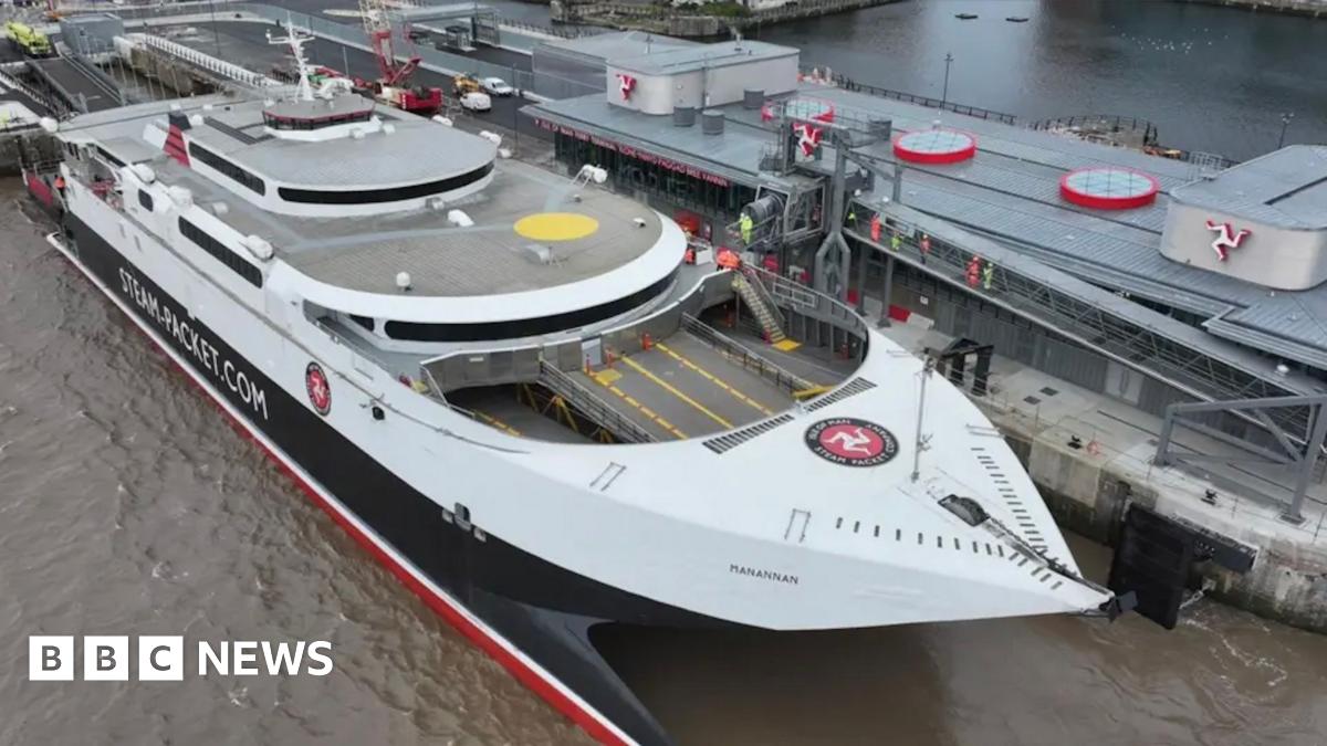 The fastcraft Manannan Steam Packet Company vessel in white and black, is docked alongside a long building with the Manx triskelion displayed on the left, right and centre of the building. People in high vis jackets can been seen on an elevated walkway connecting the ship to the land.