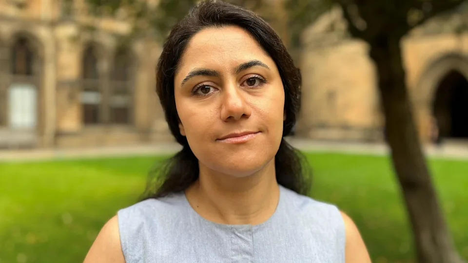 Prof Basiri looks at the camera. She is outside in the quadrangle at Glasgow university. The university buildings can be seen in the background. She has dark hair and is wearing a blue sleeveless tunic top.