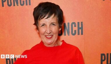 Julie Hesmondhalgh attends the press night after party for "Punch". She is wearing a red outfit, red lipstick and has short dark brown hair. She is smiling in front of an orange background with the words "punch" written in black.