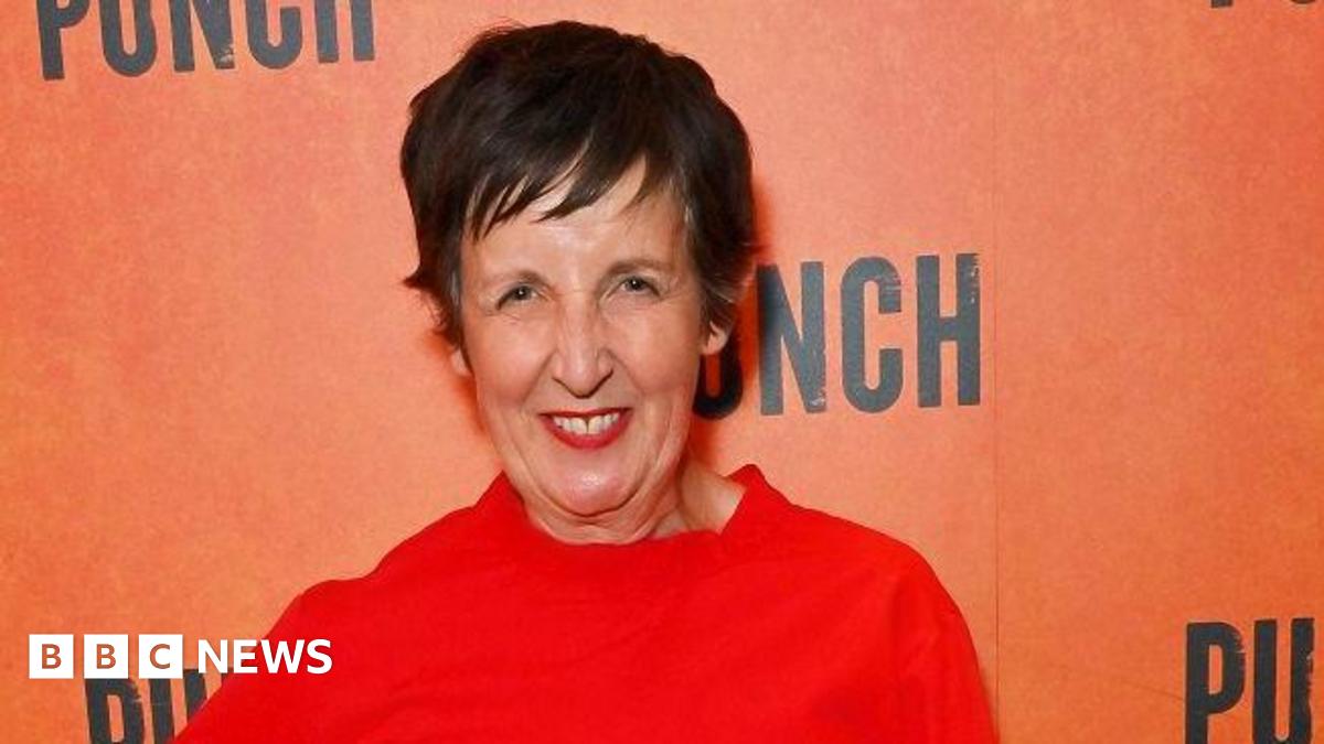 Julie Hesmondhalgh attends the press night after party for "Punch". She is wearing a red outfit, red lipstick and has short dark brown hair. She is smiling in front of an orange background with the words "punch" written in black.