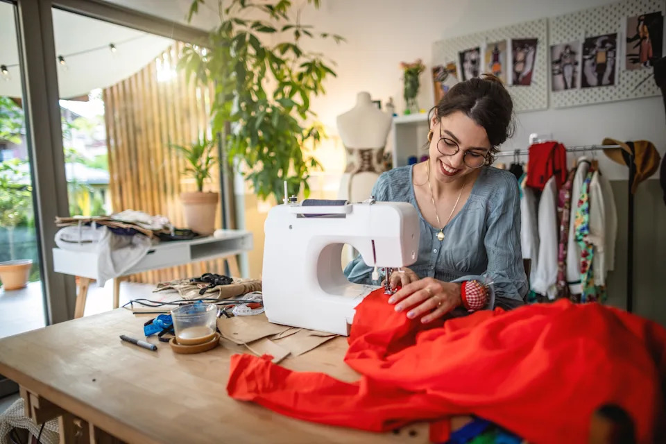 Person sewing at a table with a sewing machine, surrounded by fabric and materials in a cozy, well-lit room