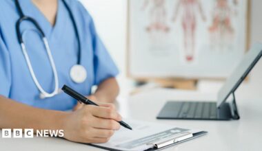 A nurse is writing on a clipboard with a black pen. There is a laptop in front of the nurse. On the table is a laptop. The nurse has blue overalls on. The background is blurred but there is a chart on display.