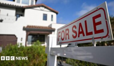 A white sign reading 'for sale' in red letters hangs in front of a white house.
