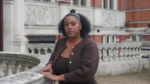 Katia Pirnak Nadine is standing outside, looking at the camera. She has black curly hair and is wearing a brown cardigan.