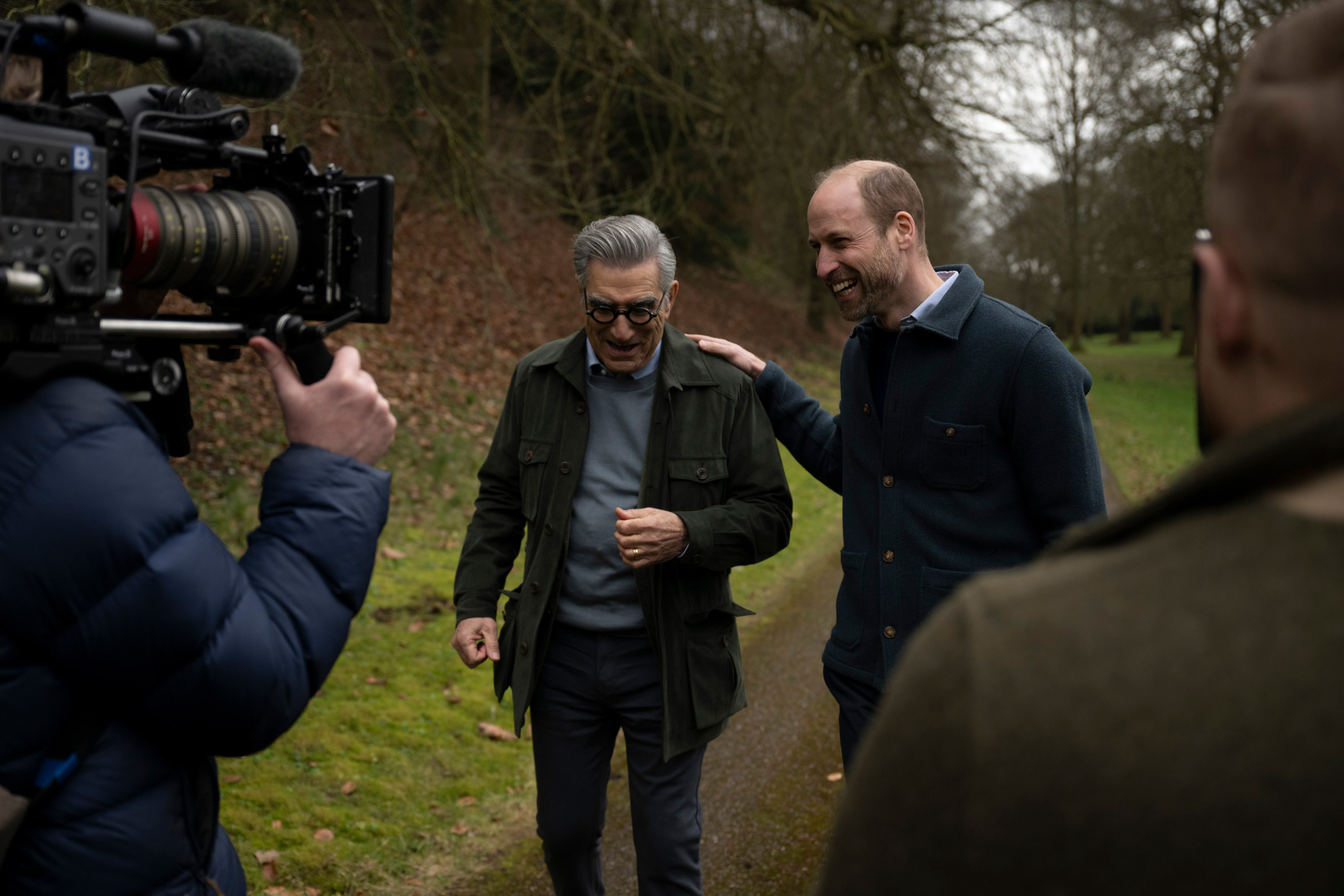 Prince William putting his hand on Eugene Levy's shoulder walking in the park while a cameraman films him