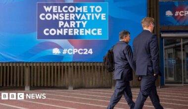 Two men in suits walk past a huge poster at the Conservative Party Conference in Manchester. The poster reads: welcome to conservative party conference.