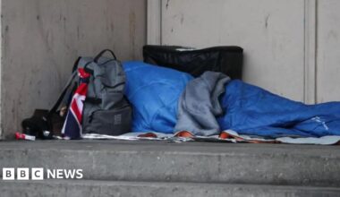 An unidentified homeless person sleeping rough in a blue sleeping bag in a doorway. A grey rucksack is in front of the person.