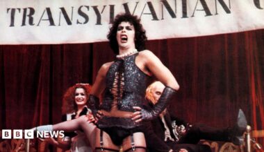 Tim Curry in a scene from the Rocky Horror Picture Show, wearing a black corset and stockings. He is singing in front of a Transylvania banner as other cast members watch