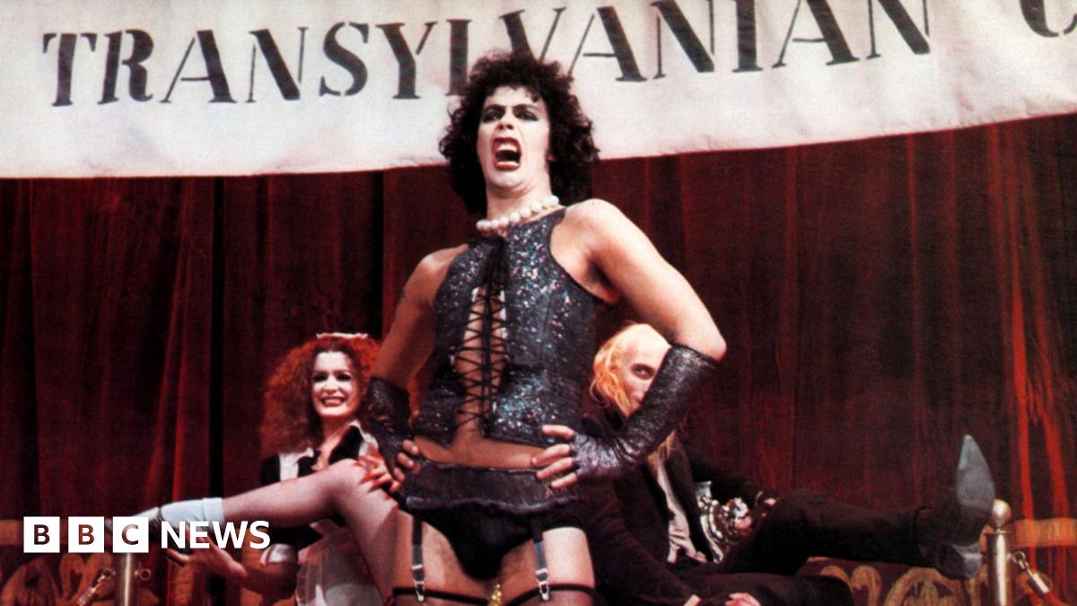Tim Curry in a scene from the Rocky Horror Picture Show, wearing a black corset and stockings. He is singing in front of a Transylvania banner as other cast members watch