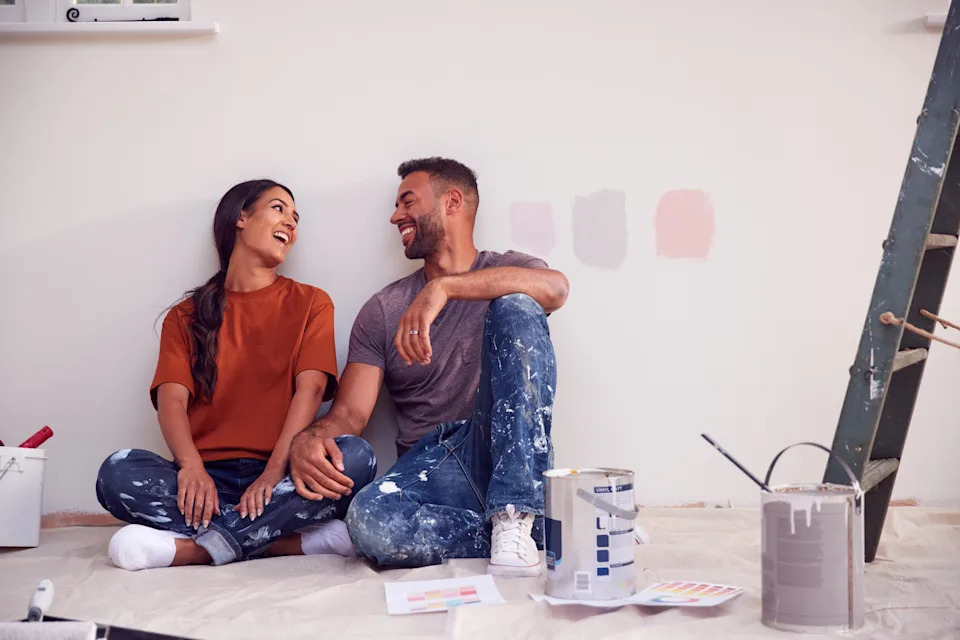 A couple sits on the floor, smiling at each other, surrounded by paint cans and swatches, likely planning a room painting project