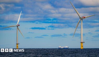 A ship passing between two offshore wind turbines on a dark blue sea under a light blue sky with grey and white clouds.