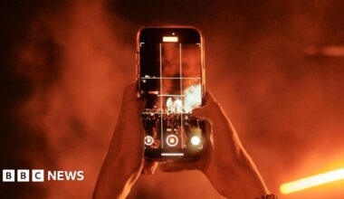 Somebody holding a phone up in a dark venue. Orange lights can be seen in the distance. The person is filming on their phone.