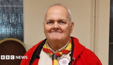 Matthew Padley with white hair wearing a Scout leader uniform complete with red fleece beige shirt and red and yellow necker. He is smiling.